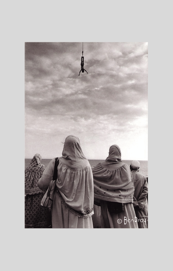 A girl dangles into the top of the picture, upsidedown, arms spread wide, while a group of women wearing headscarves watch.