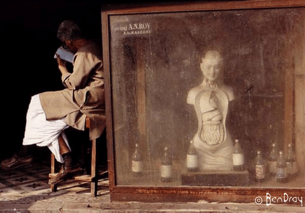 An old man and is sitting next to an anatomical model. The model is advertising a pharmacy in Delhi, India.
