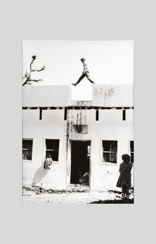 A boy in Pushkar Rajasthan steps over the roofs around a courtyard while a girl watches. This is a black and white portrait photo.