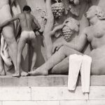 Black and white image of a semi naked man climbing across a frieze of statues.